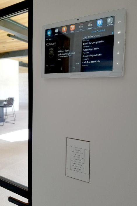 Wall-mounted touchscreen displaying home automation controls and music playlist, with a patio visible through a glass door