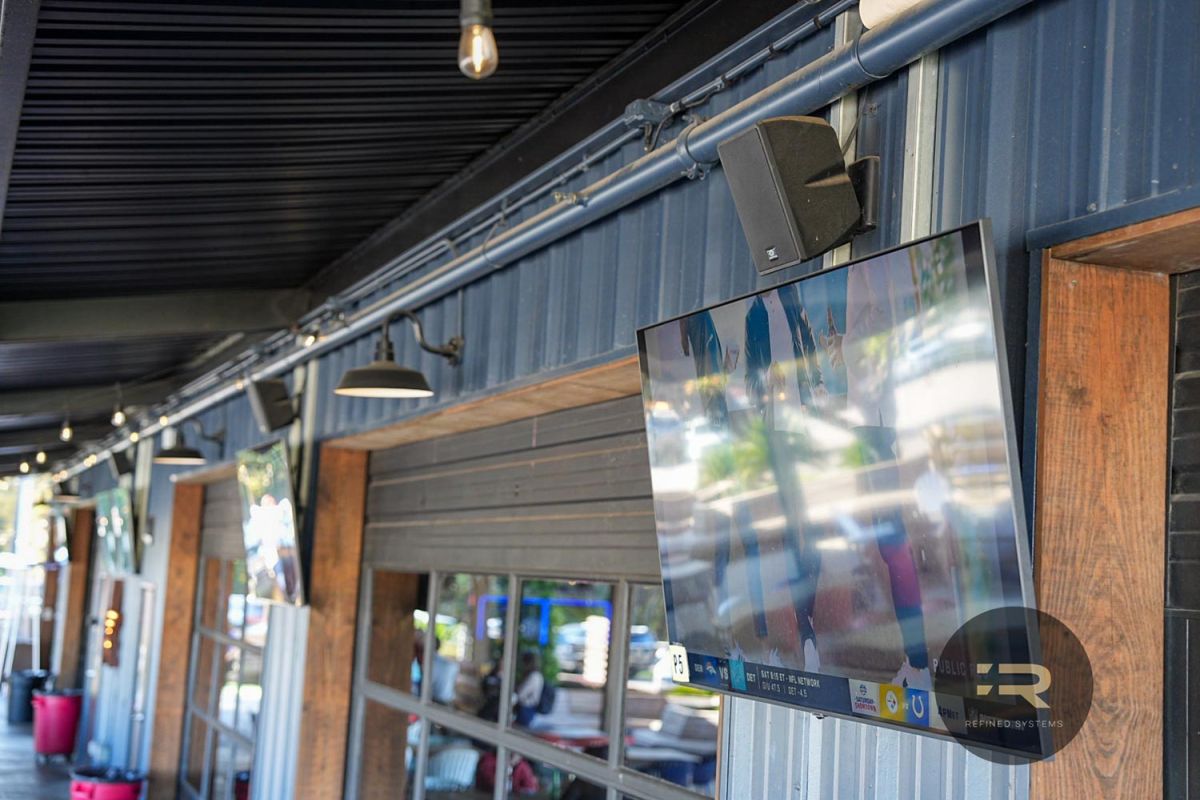 Multiple outdoor TVs mounted along a patio wall with black fixtures and speakers.
