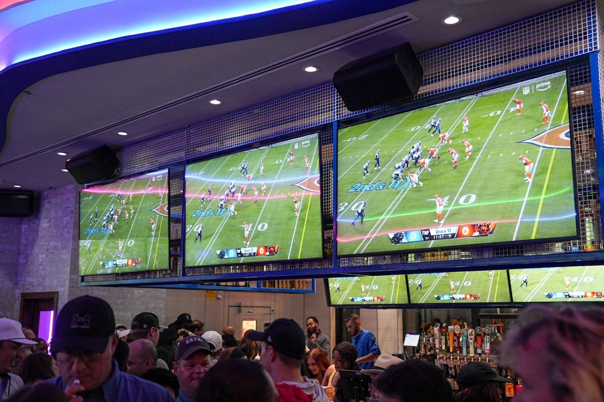 Three large TVs displaying a football play above a busy bar counter.