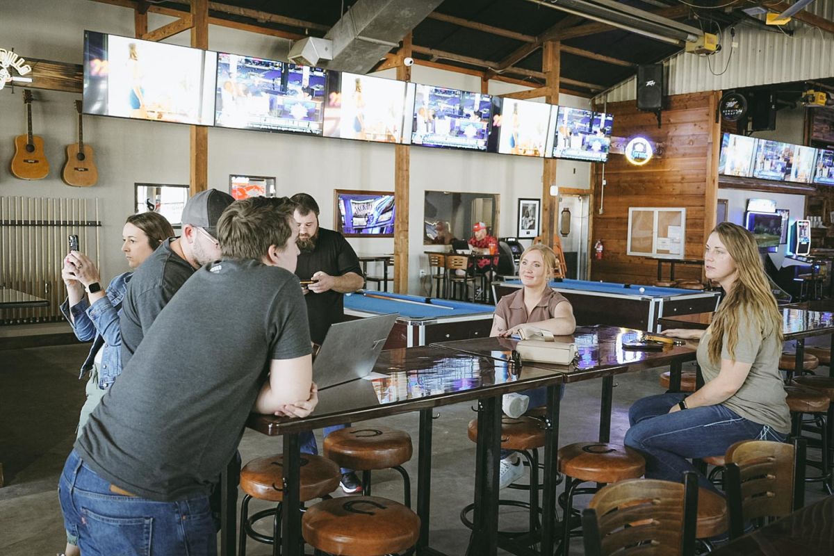 Group of people chatting around a tall table under multiple TV screens