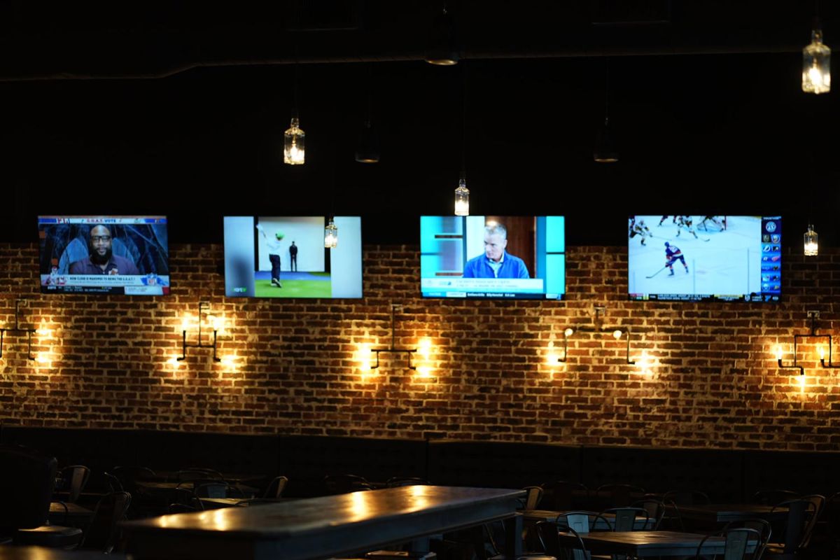 Row of four TVs mounted on a brick wall, each displaying different sports or shows.