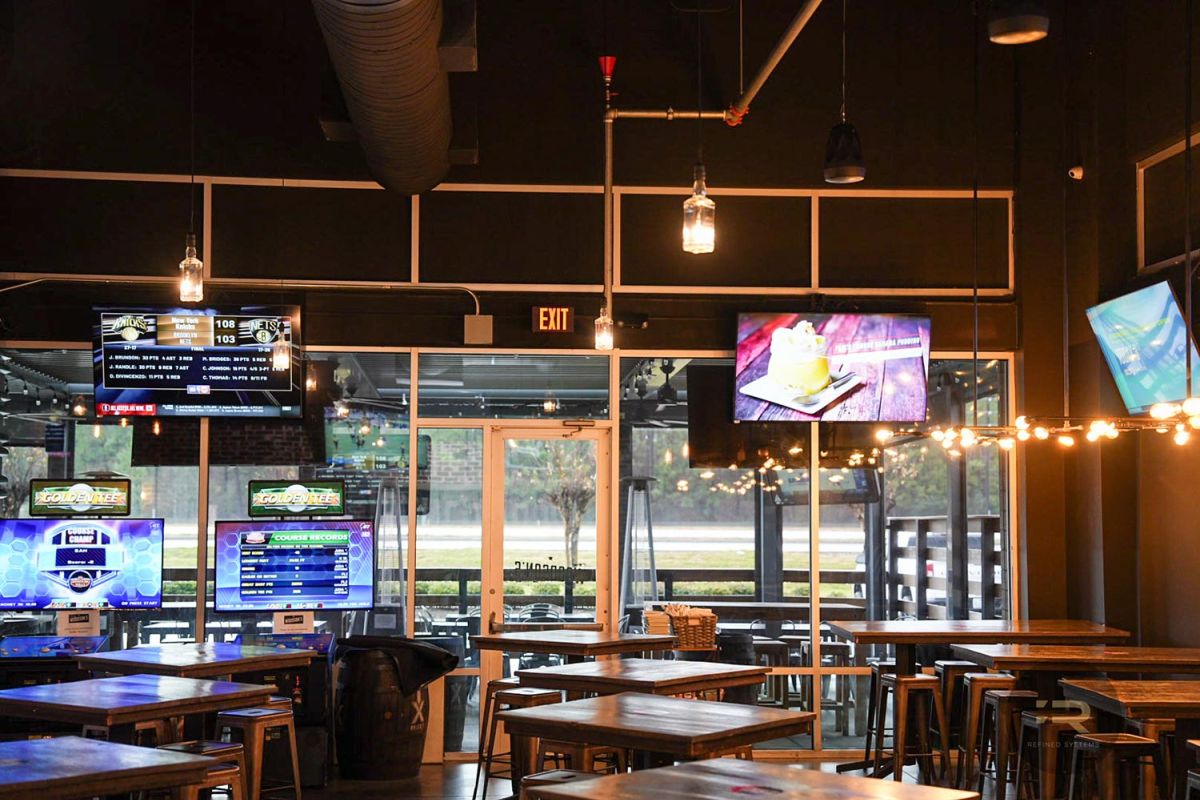 TVs and arcade machines near a glass exit with string lights and wooden tables.