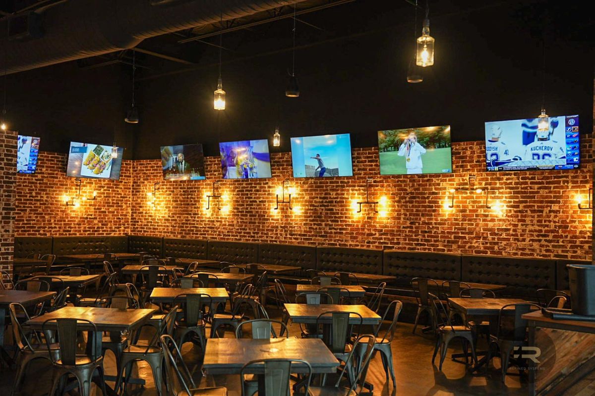 Corner of a bar with brick walls and multiple screens showing food and sports.